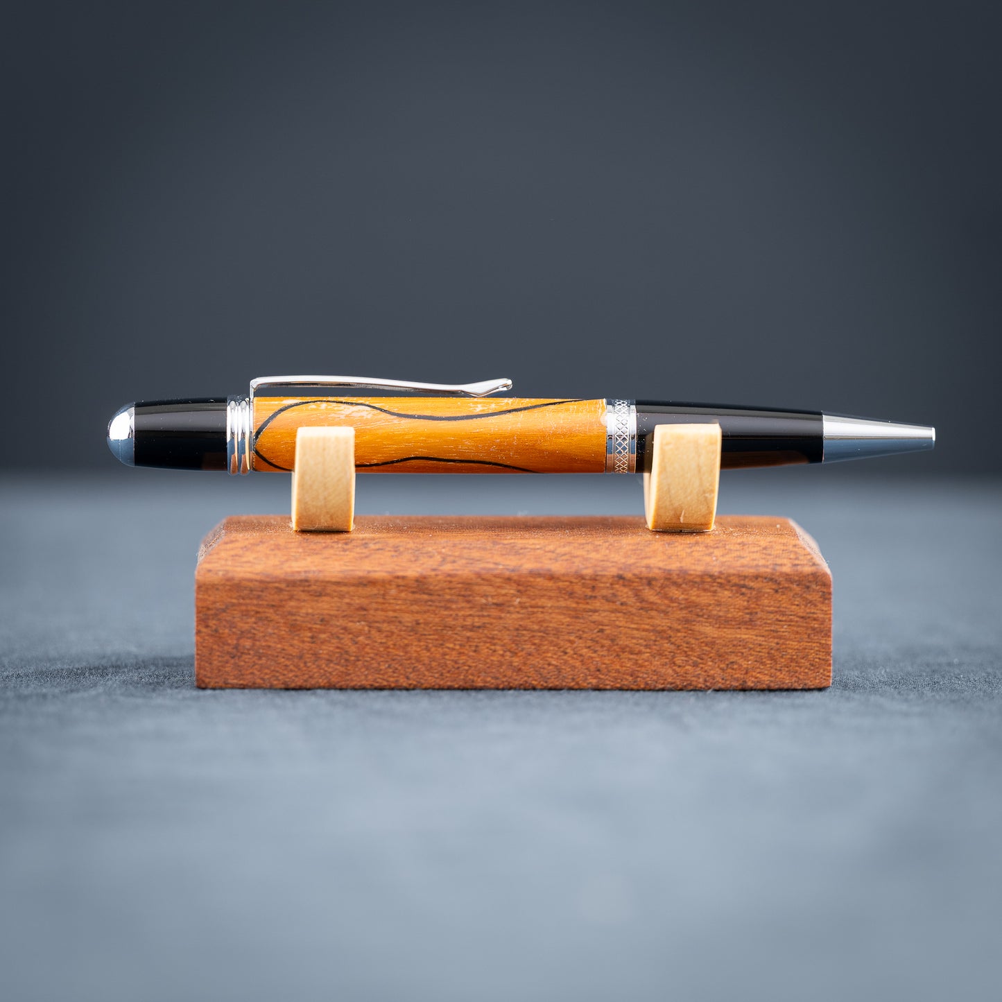 Handmade Osage orange wood and black wood ballpoint pen on a black stand against a wood background.