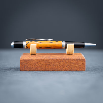 Handmade Osage orange wood and black wood ballpoint pen on a black stand against a wood background.