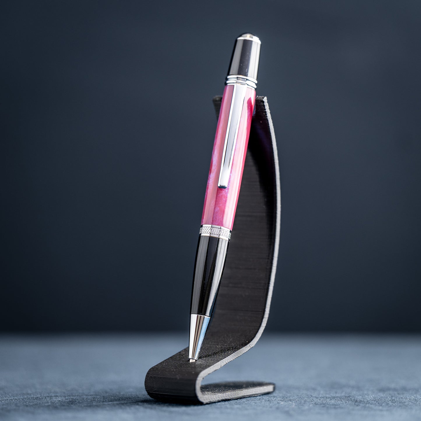 Handmade pink, purple, and blue resin twist ballpoint pen with black and chrome details on a dark stand against a dark background.