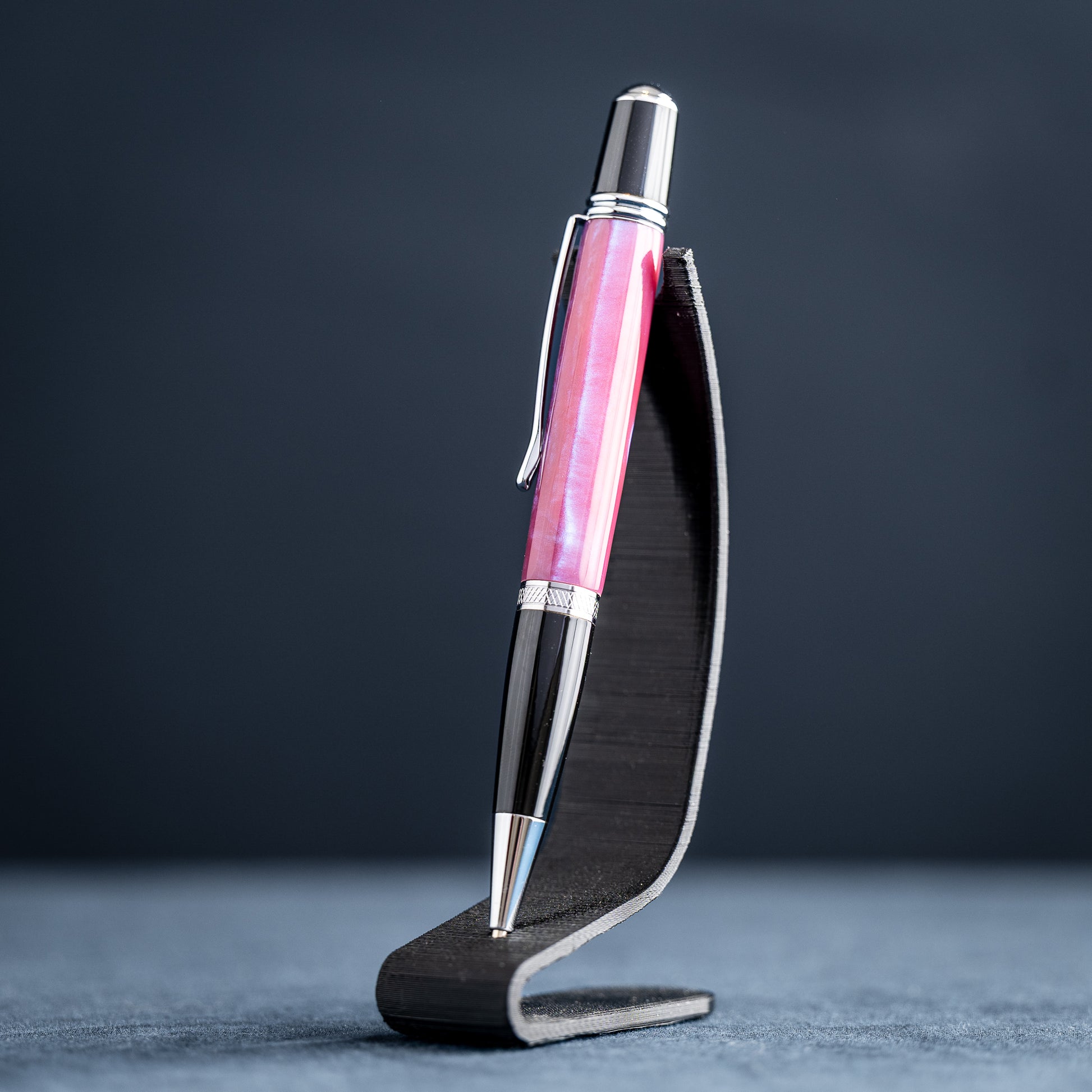 Handmade pink, purple, and blue resin twist ballpoint pen with black and chrome details on a dark stand against a dark background.