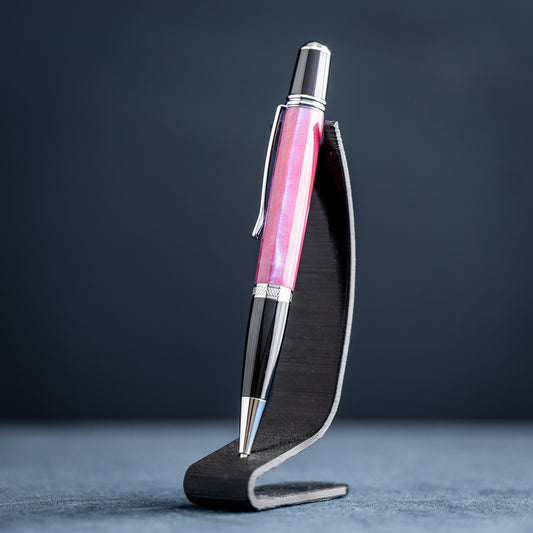 Handmade pink, purple, and blue resin twist ballpoint pen with black and chrome details on a dark stand against a dark background.