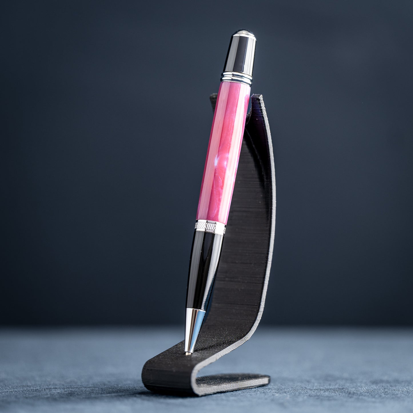 Handmade pink, purple, and blue resin twist ballpoint pen with black and chrome details on a dark stand against a dark background.