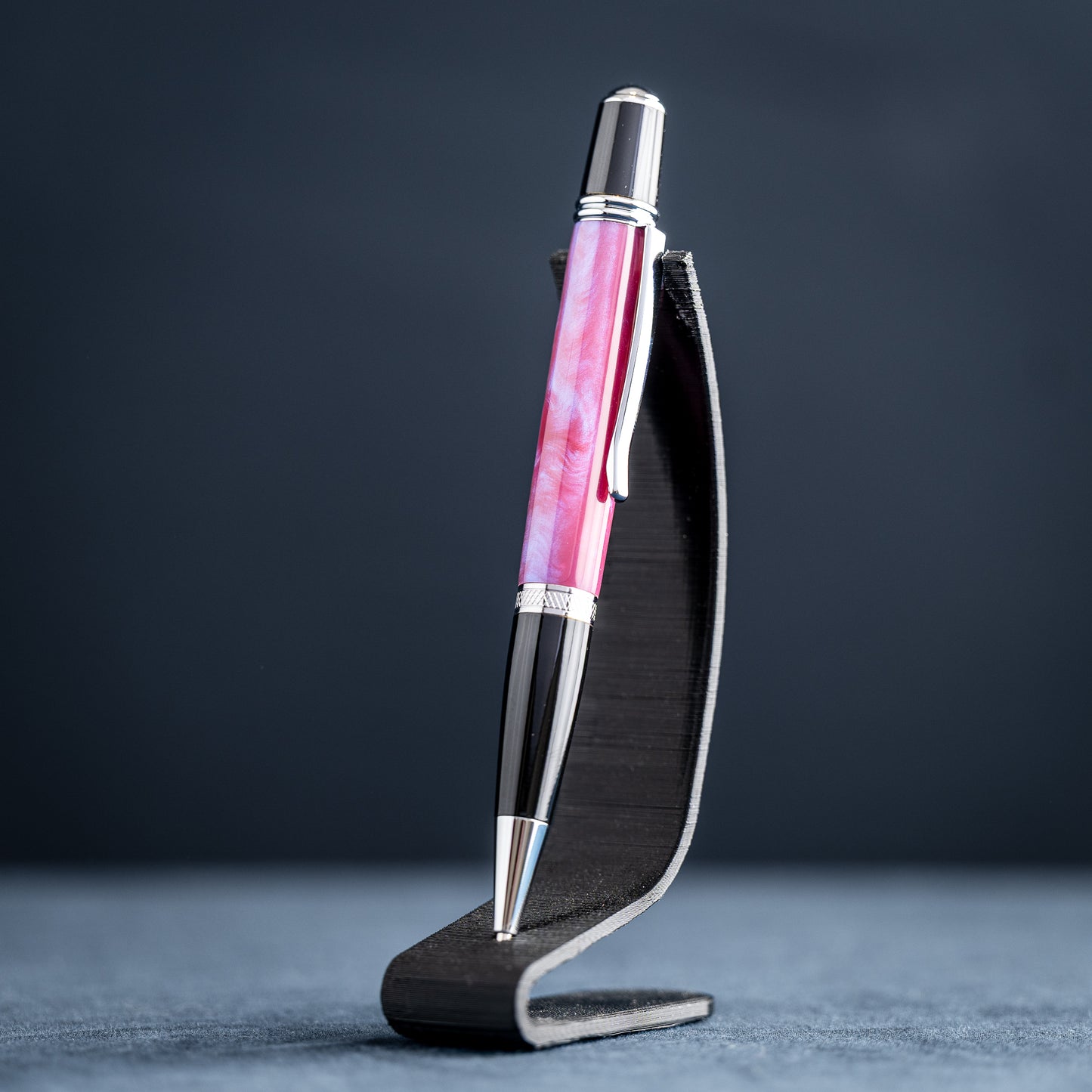 Handmade pink, purple, and blue resin twist ballpoint pen with black and chrome details on a dark stand against a dark background.