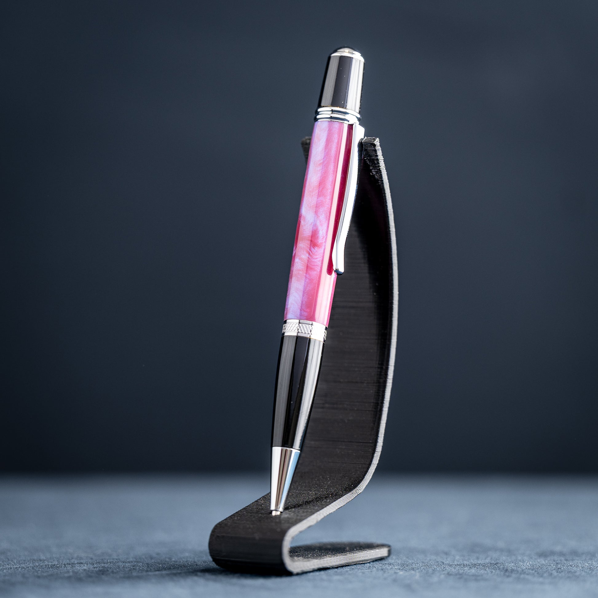 Handmade pink, purple, and blue resin twist ballpoint pen with black and chrome details on a dark stand against a dark background.