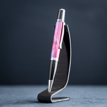 Handmade pink, purple, and blue resin twist ballpoint pen with black and chrome details on a dark stand against a dark background.