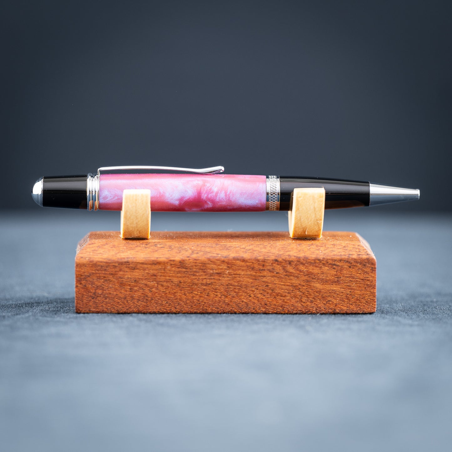 Handmade pink, purple, and blue resin twist ballpoint pen with black and chrome details on a wooden stand against a dark background.
