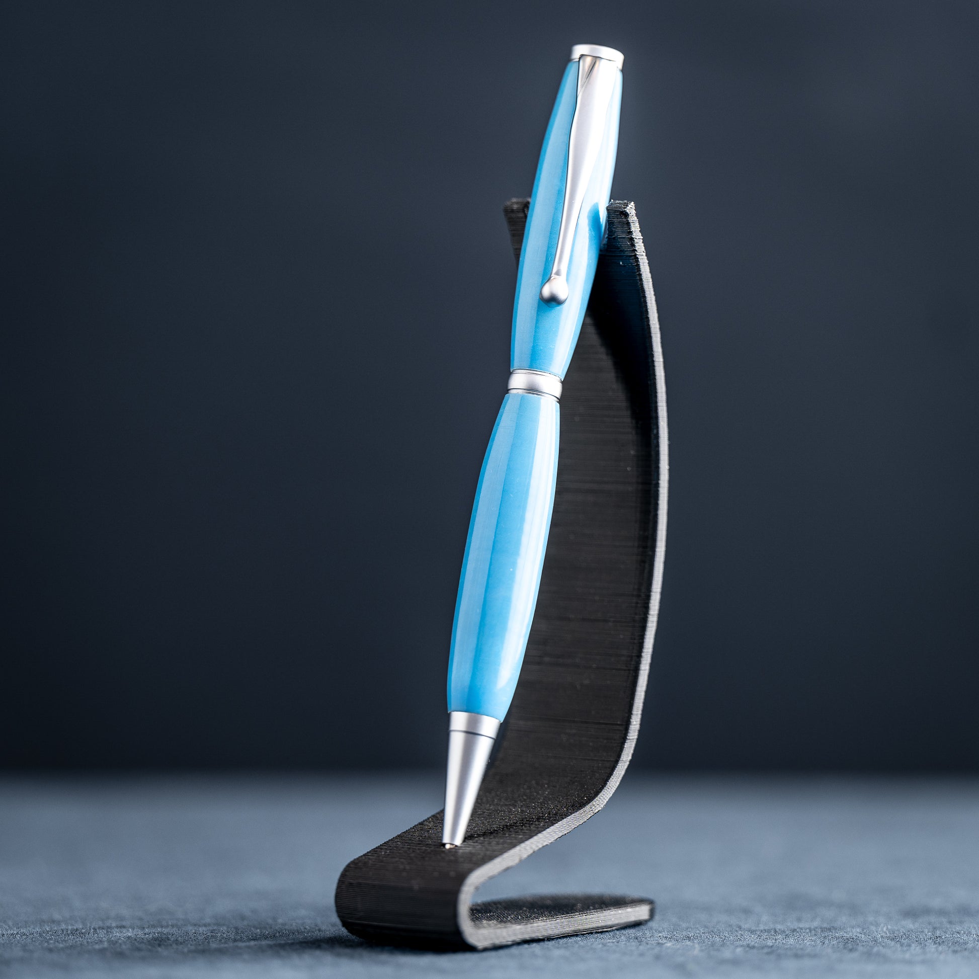 A handmade powder blue ballpoint resin twist pen with satin chrome plating on a dark stand against a dark background.