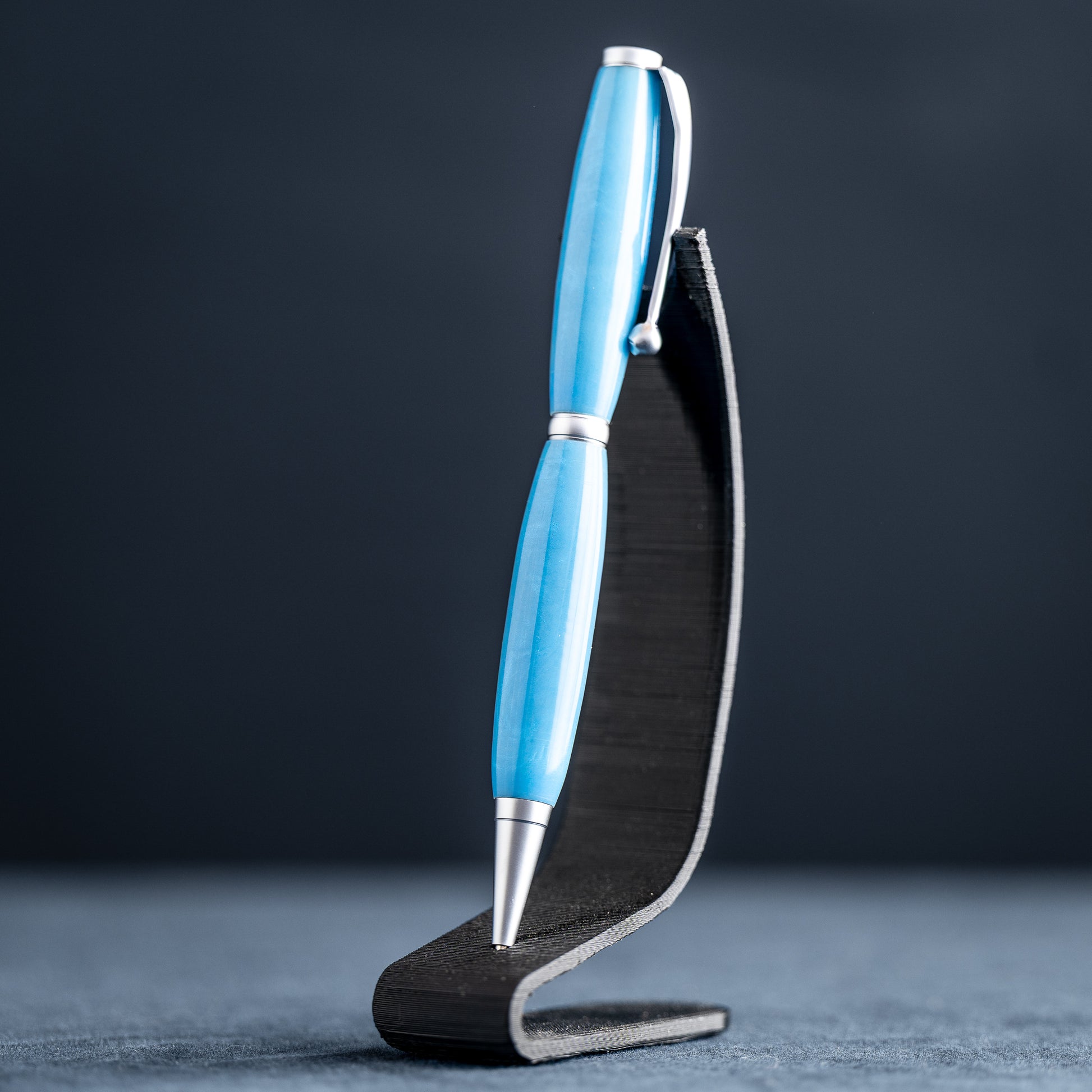 A handmade powder blue ballpoint resin twist pen with satin chrome plating on a dark stand against a dark background.