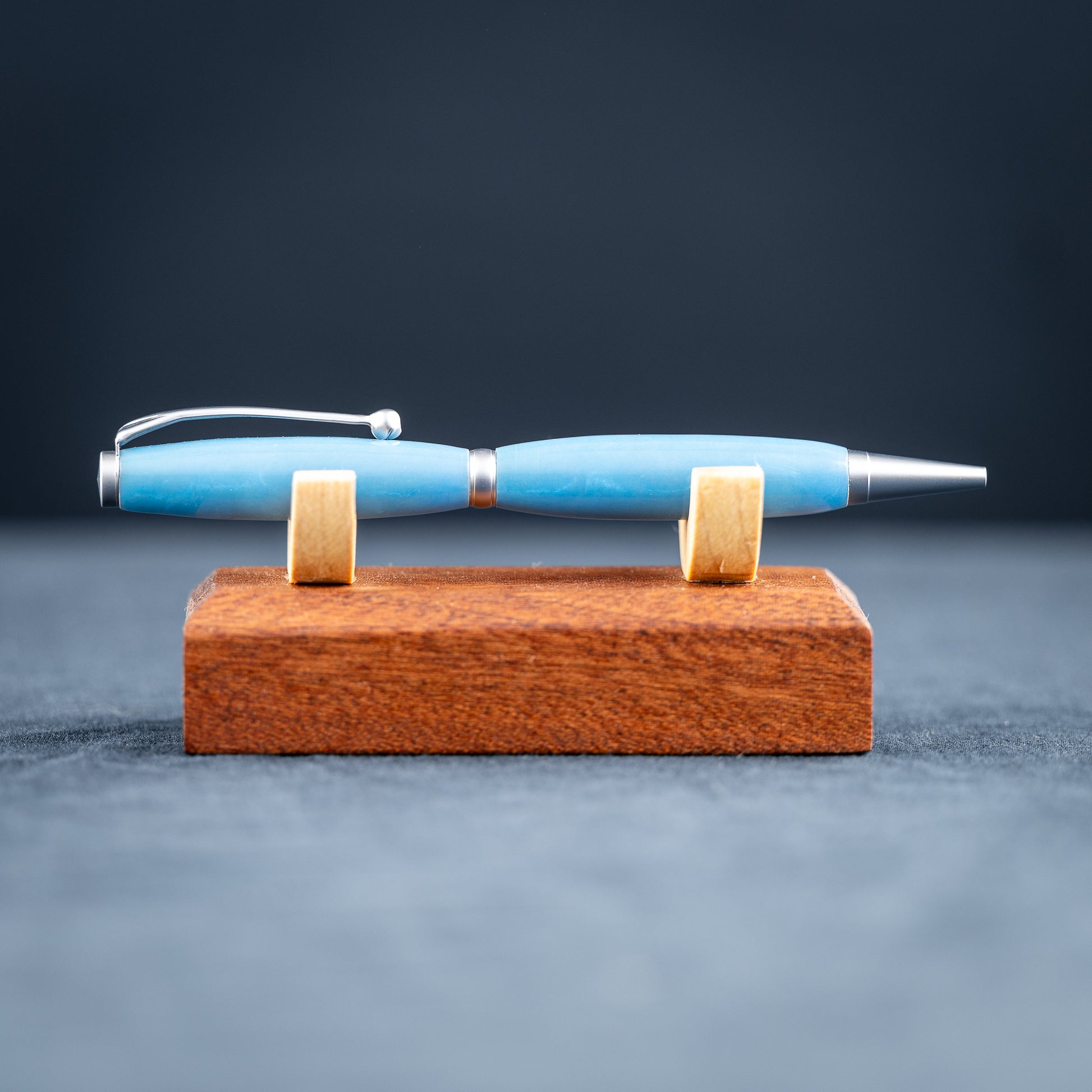 A handmade powder blue ballpoint resin twist pen with satin chrome plating on a wooden stand against a dark background.