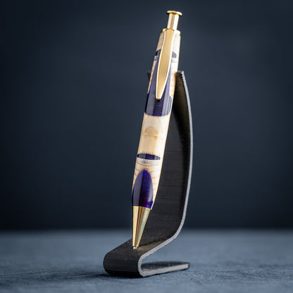 A handmade pine wood, purple, and white resin ballpoint click pen with gold plating on a wooden stand against a dark background.