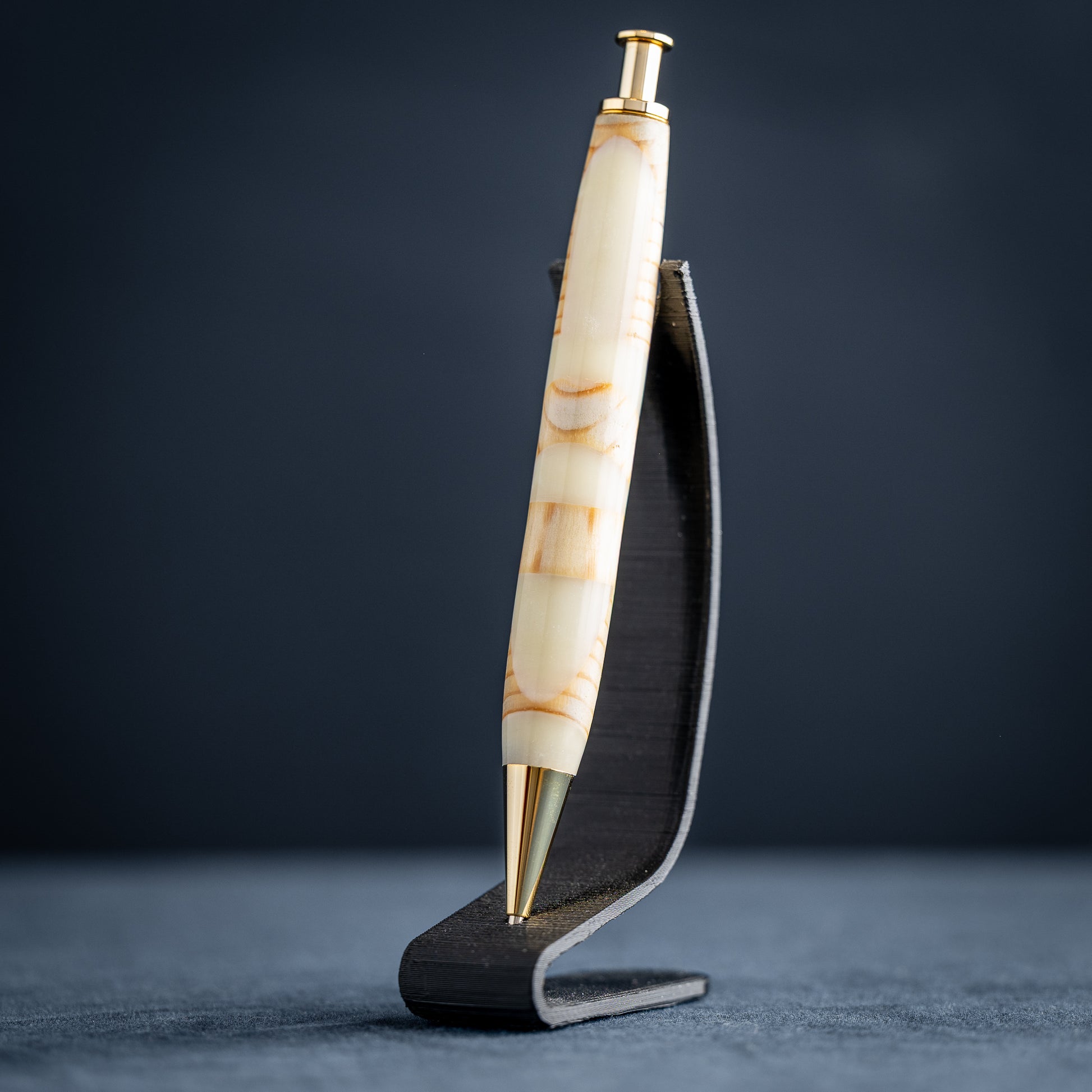 A handmade pine wood, purple, and white resin ballpoint click pen with gold plating on a wooden stand against a dark background.