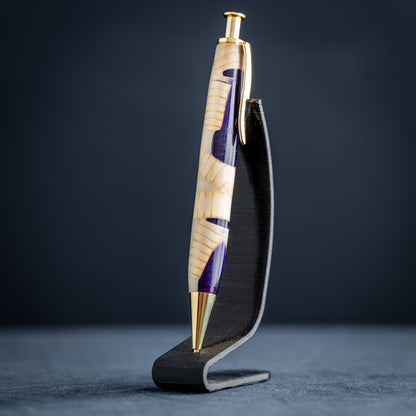 A handmade pine wood, purple, and white resin ballpoint click pen with gold plating on a wooden stand against a dark background.