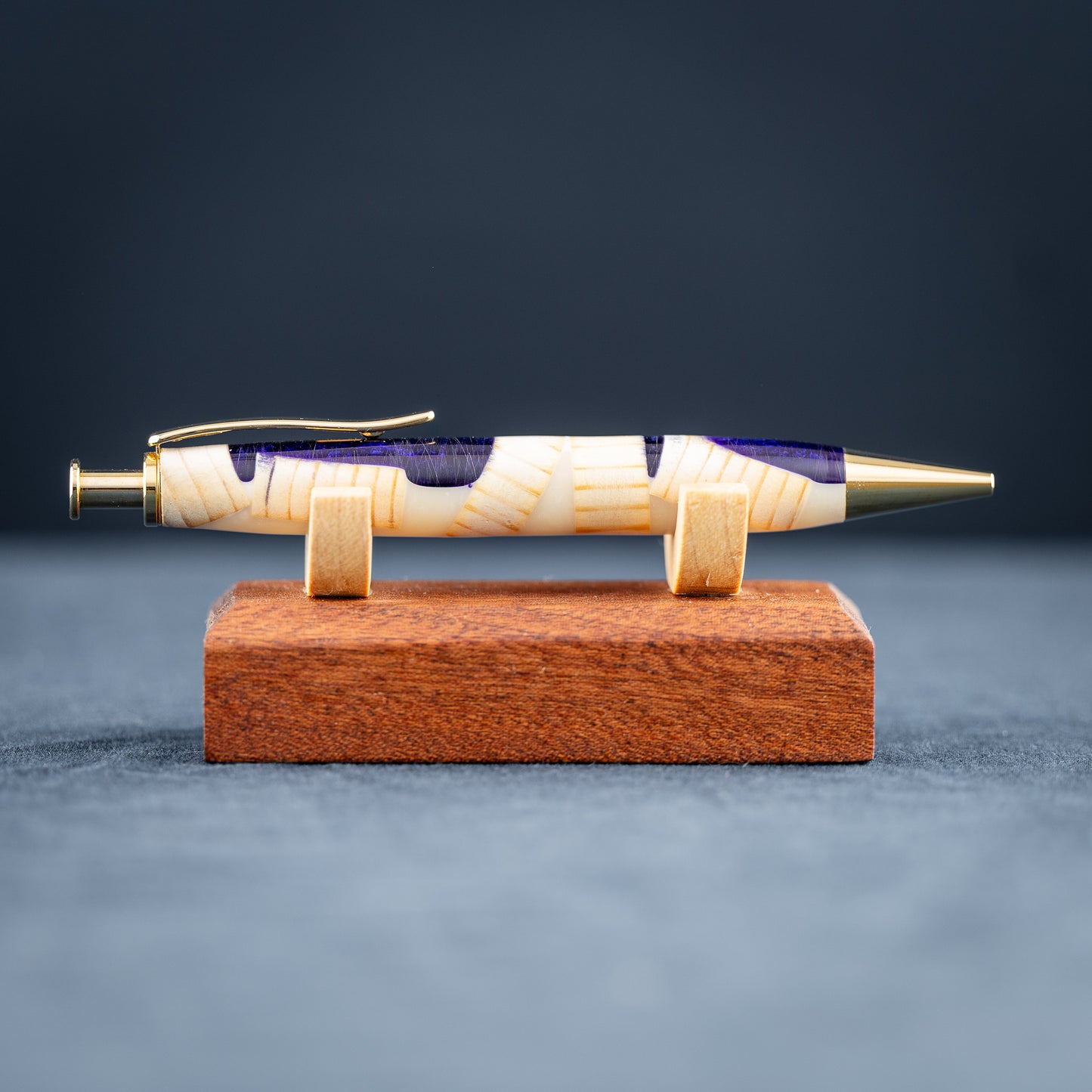 A handmade pine wood, purple, and white resin ballpoint click pen with gold plating on a wooden stand against a dark background.