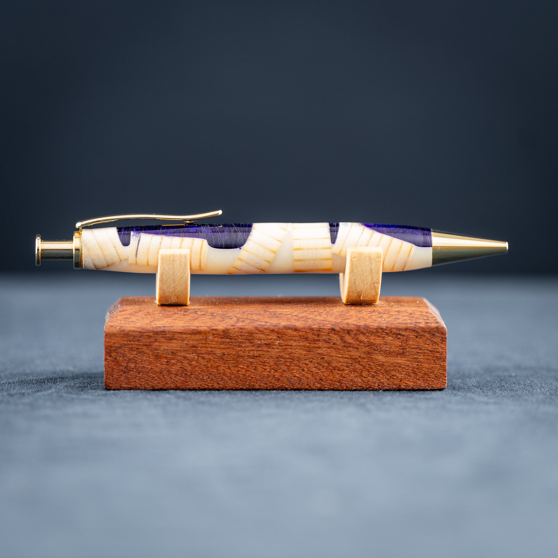 A handmade pine wood, purple, and white resin ballpoint click pen with gold plating on a wooden stand against a dark background.