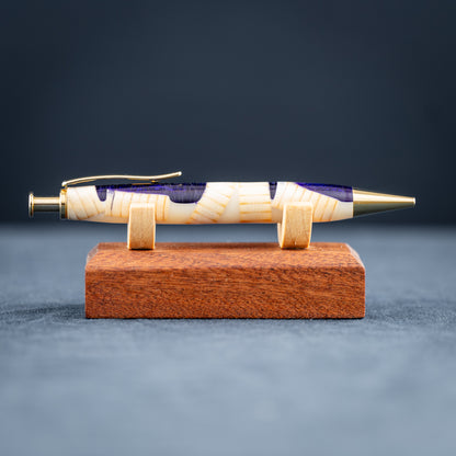 A handmade pine wood, purple, and white resin ballpoint click pen with gold plating on a wooden stand against a dark background.