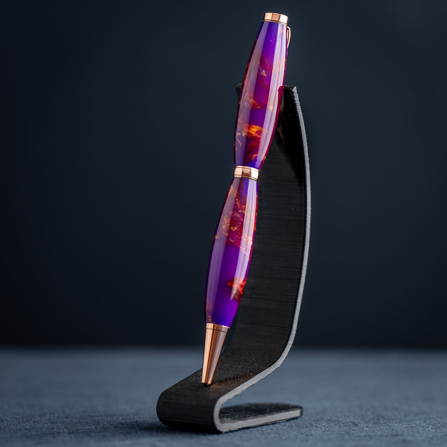 A handmade purple, bronze, and copper resin swirl ballpoint pen with copper plating on a dark stand against a dark background.