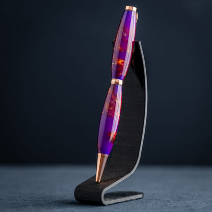A handmade purple, bronze, and copper resin swirl ballpoint pen with copper plating on a dark stand against a dark background.