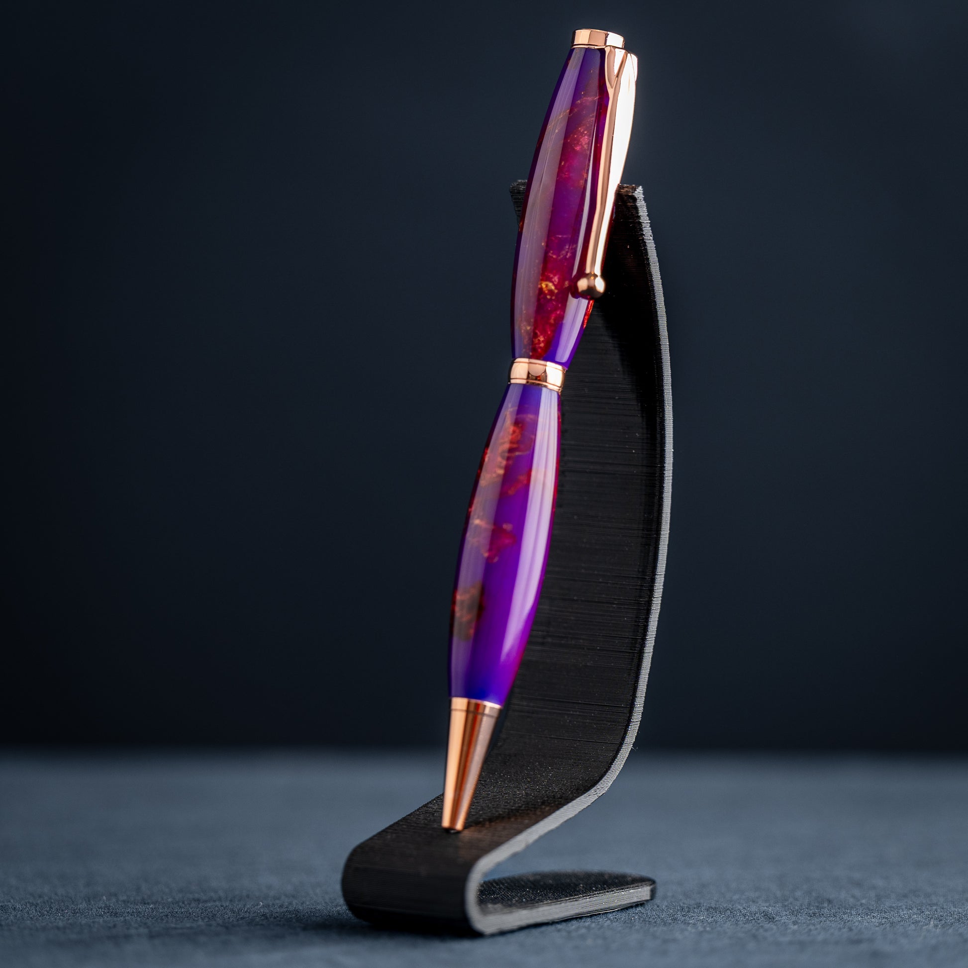 A handmade purple, bronze, and copper resin swirl ballpoint pen with copper plating on a dark stand against a dark background.