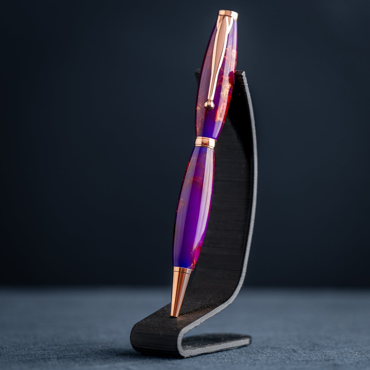 A handmade purple, bronze, and copper resin swirl ballpoint pen with copper plating on a dark stand against a dark background.