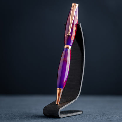 A handmade purple, bronze, and copper resin swirl ballpoint pen with copper plating on a dark stand against a dark background.