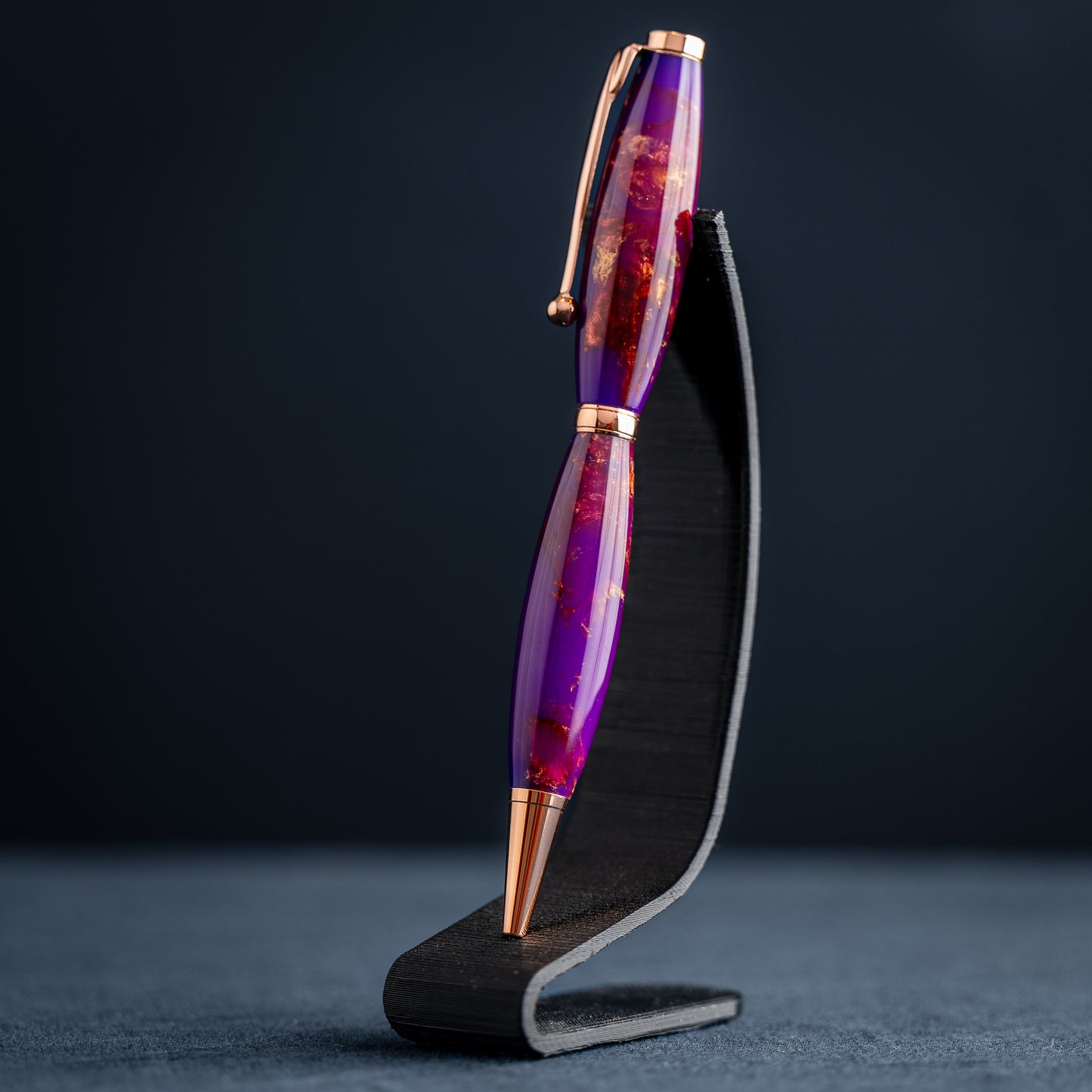 A handmade purple, bronze, and copper resin swirl ballpoint pen with copper plating on a dark stand against a dark background.
