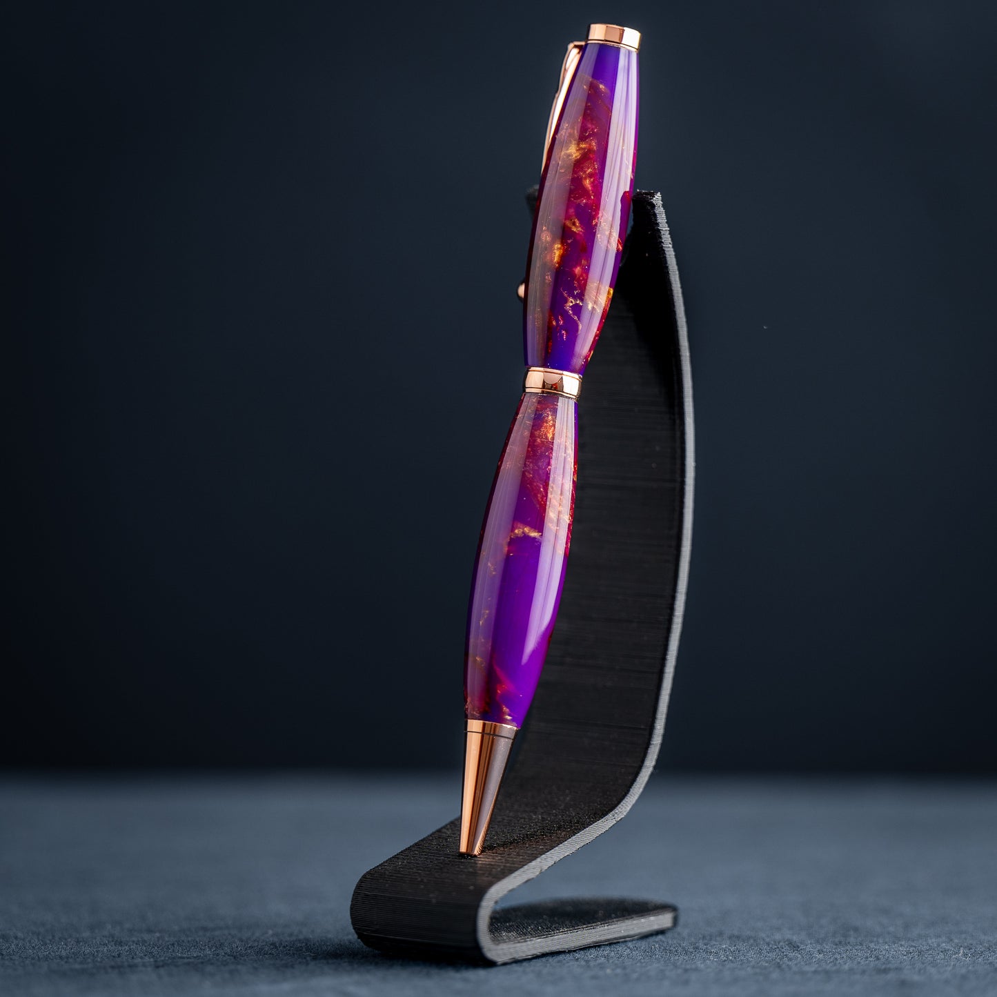 A handmade purple, bronze, and copper resin swirl ballpoint pen with copper plating on a dark stand against a dark background.