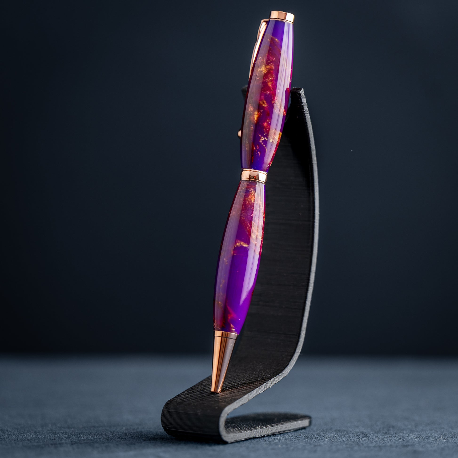 A handmade purple, bronze, and copper resin swirl ballpoint pen with copper plating on a dark stand against a dark background.