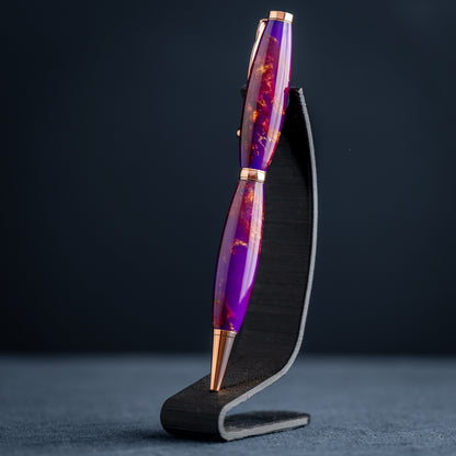 A handmade purple, bronze, and copper resin swirl ballpoint pen with copper plating on a dark stand against a dark background.