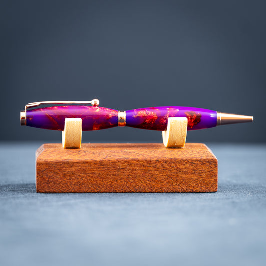 A handmade purple, bronze, and copper resin swirl ballpoint pen with copper plating on a wooden stand against a dark background.
