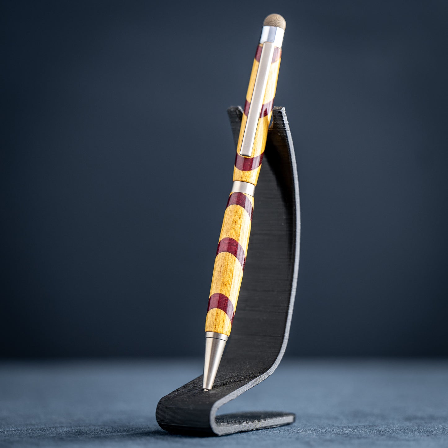 Handmade purple heart and canary wood ballpoint twist pen with stylus on a dark stand against a dark background.
