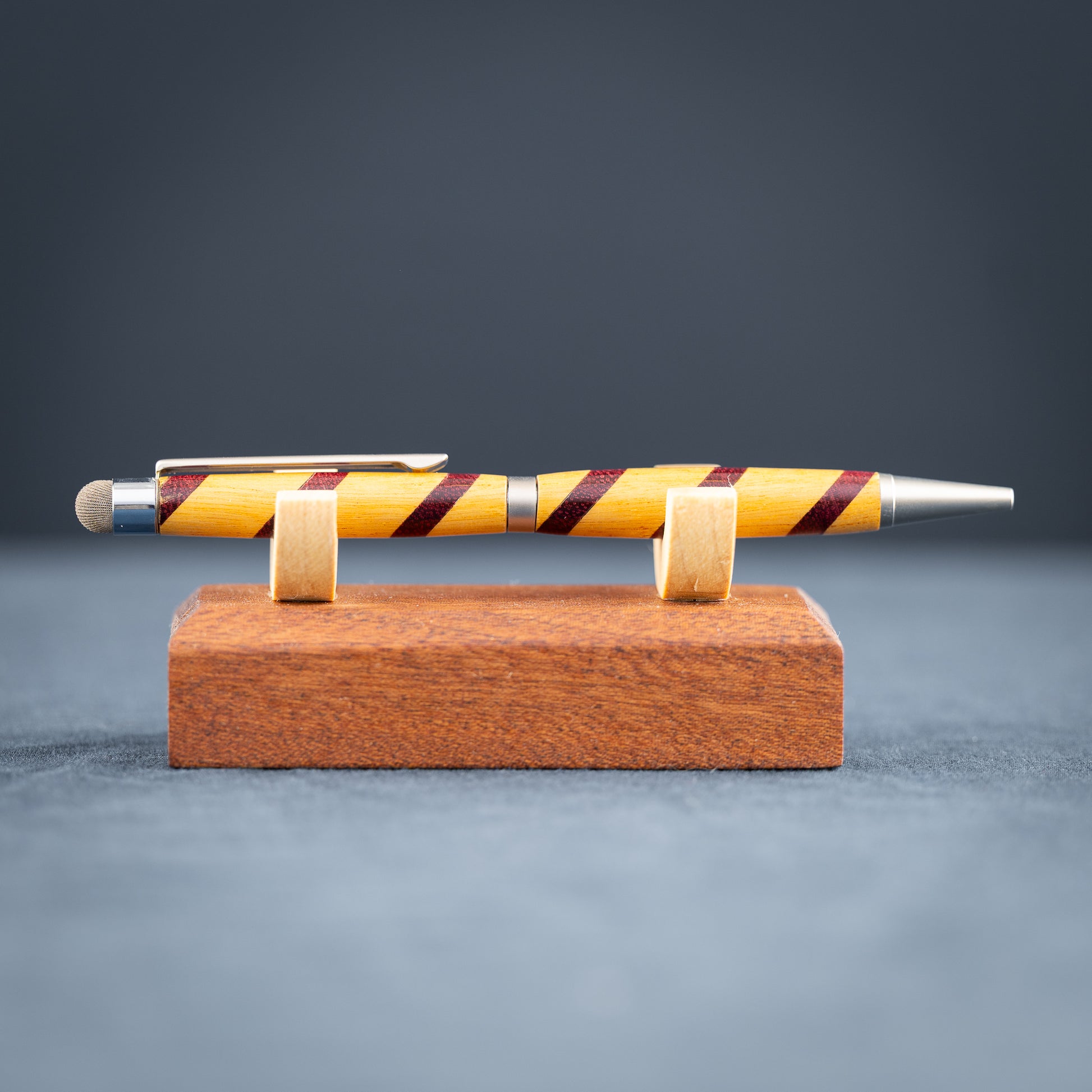 Handmade purple heart and canary wood ballpoint twist pen with stylus on a wooden stand against a dark background.