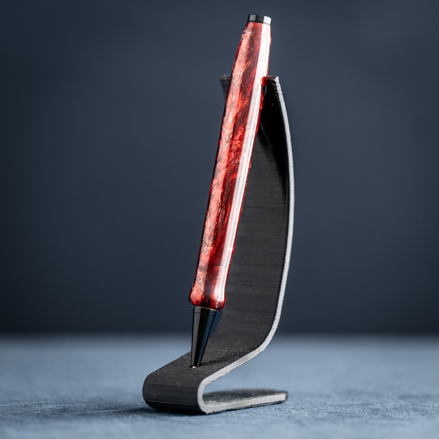 A handmade red and black resin swirl pen with black chrome plating on a dark stand against a white background.