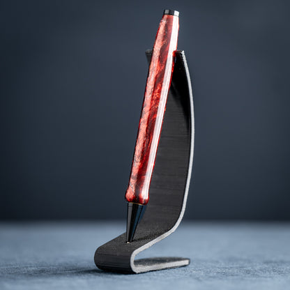 A handmade red and black resin swirl pen with black chrome plating on a dark stand against a white background.
