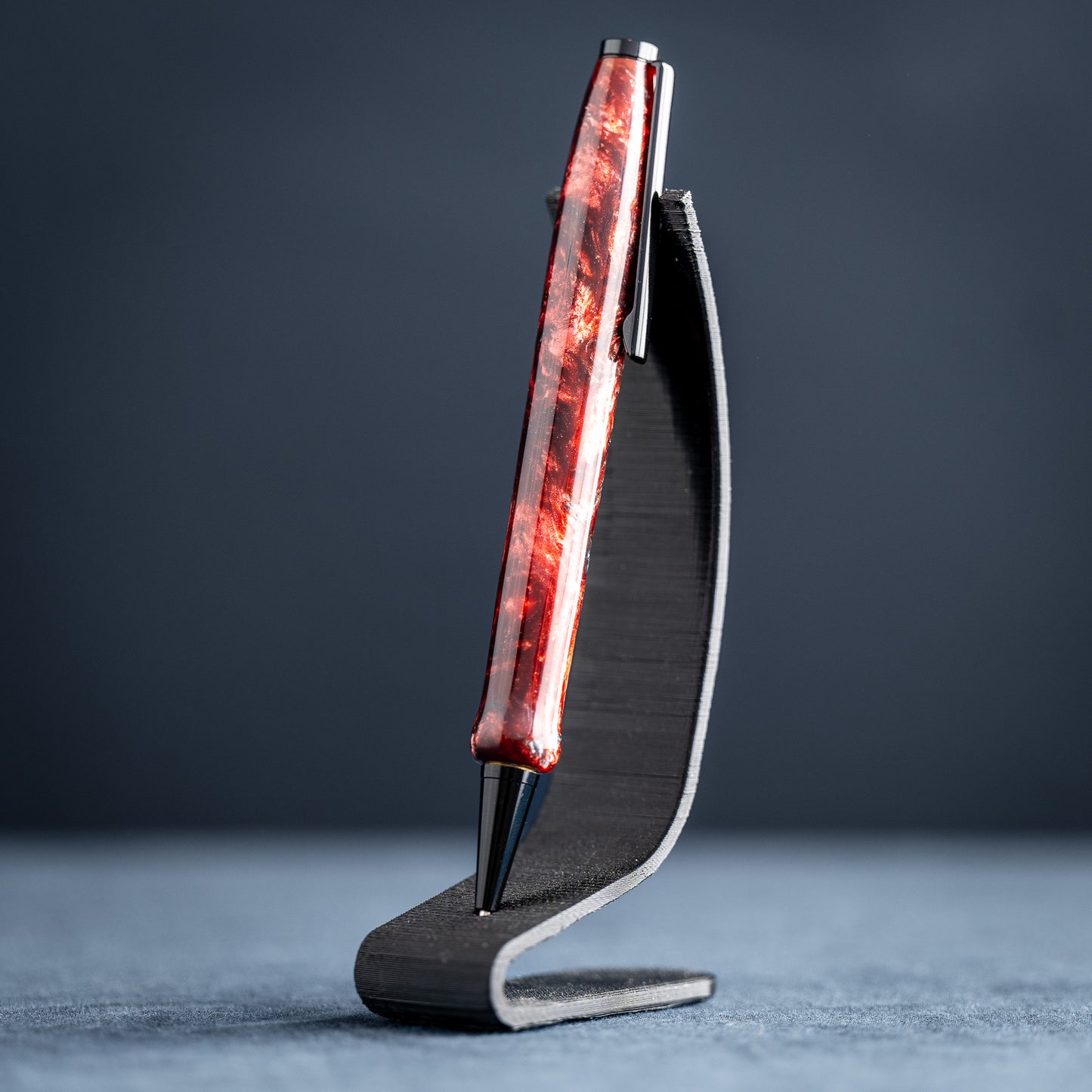 A handmade red and black resin swirl pen with black chrome plating on a dark stand against a dark background.