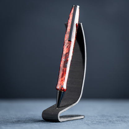 A handmade red and black resin swirl pen with black chrome plating on a dark stand against a dark background.