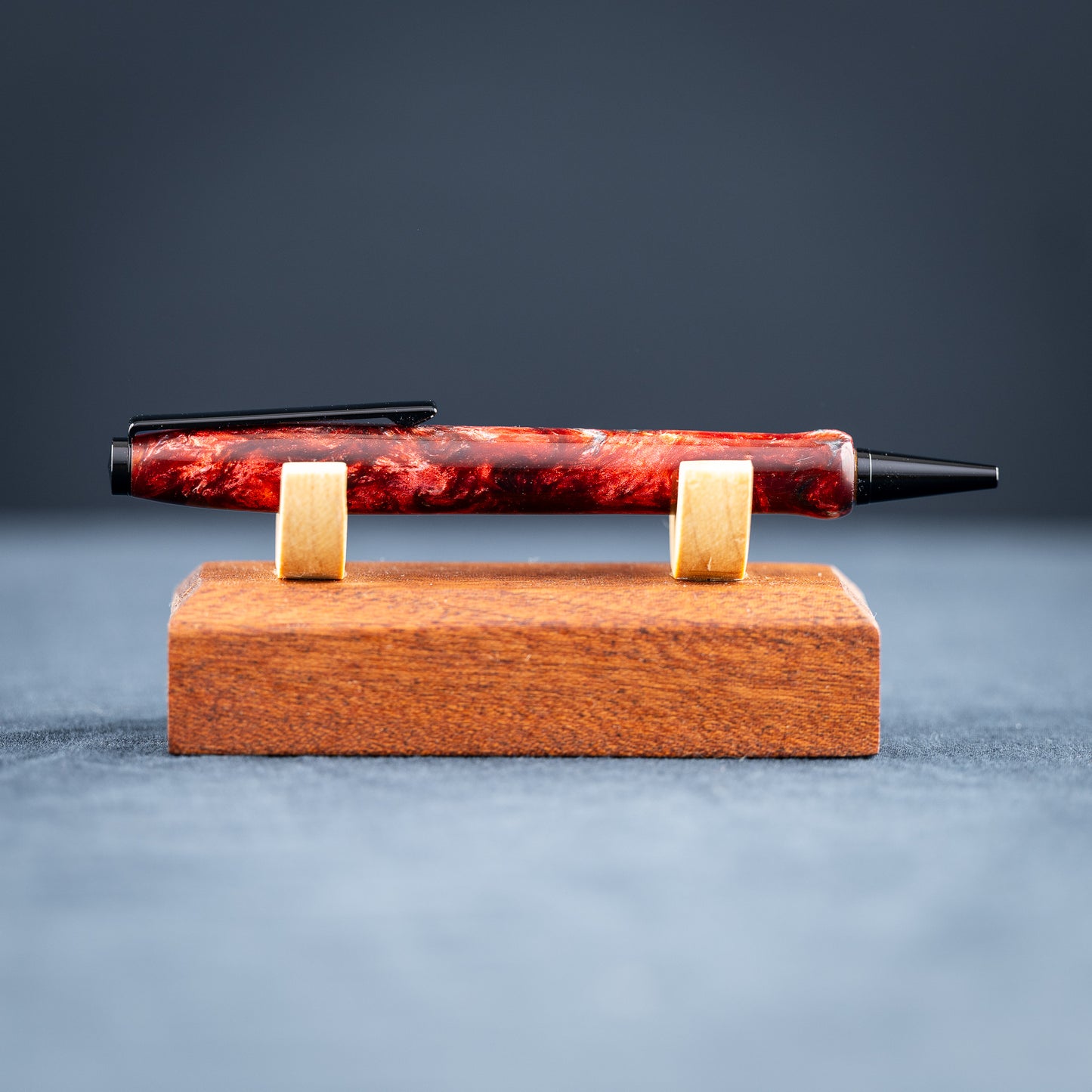 A handmade red and black resin swirl pen with black chrome plating on a dark stand against a wooden background.