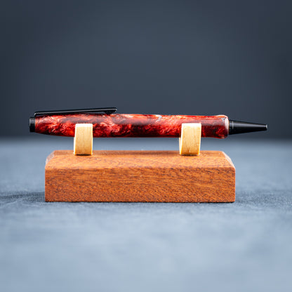 A handmade red and black resin swirl pen with black chrome plating on a dark stand against a wooden background.