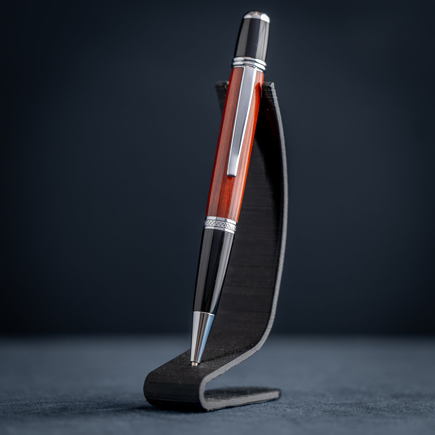 Handmade Redheart wooden ballpoint pen on a dark stand against a black background.