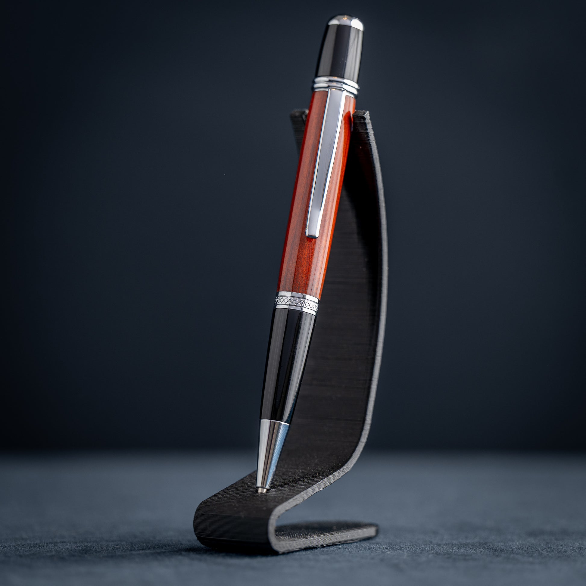 Handmade Redheart wooden ballpoint pen on a dark stand against a black background.