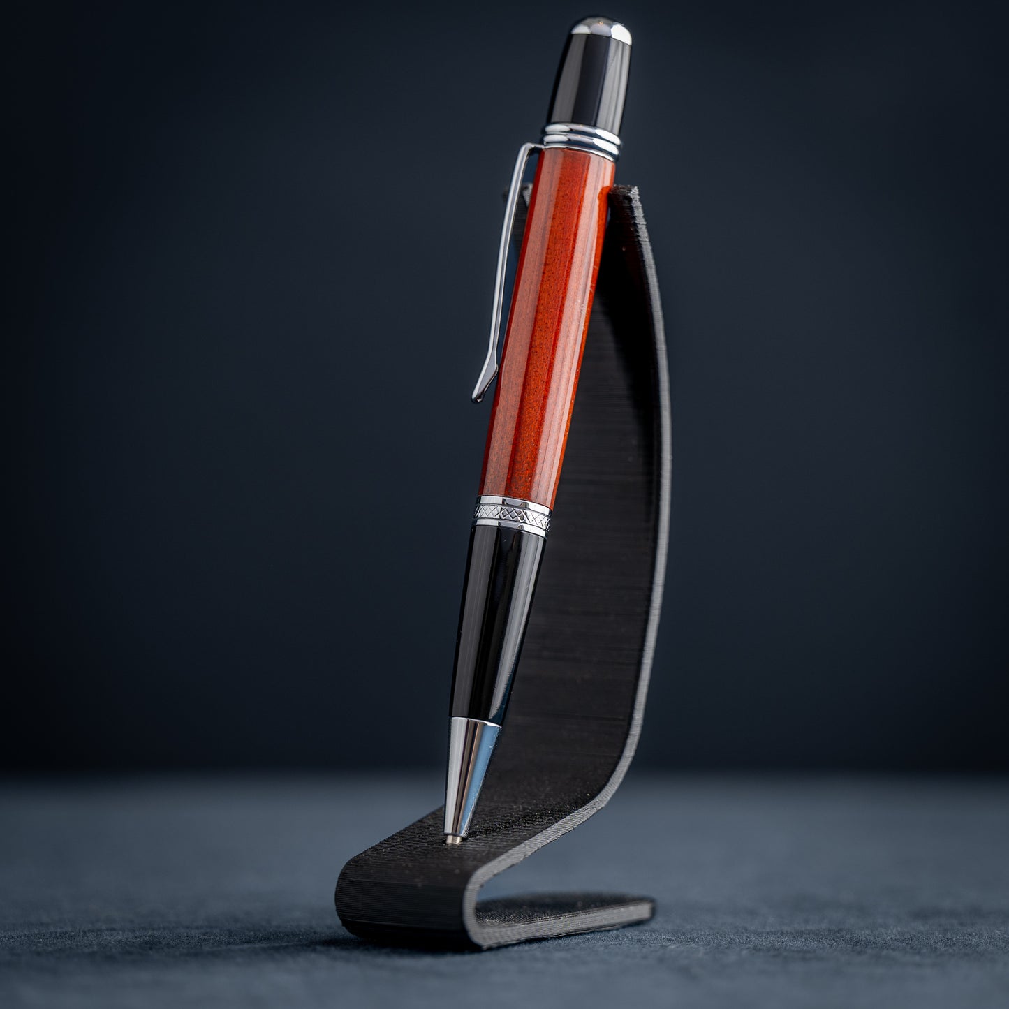 Handmade Redheart wooden ballpoint pen on a dark stand against a black background.