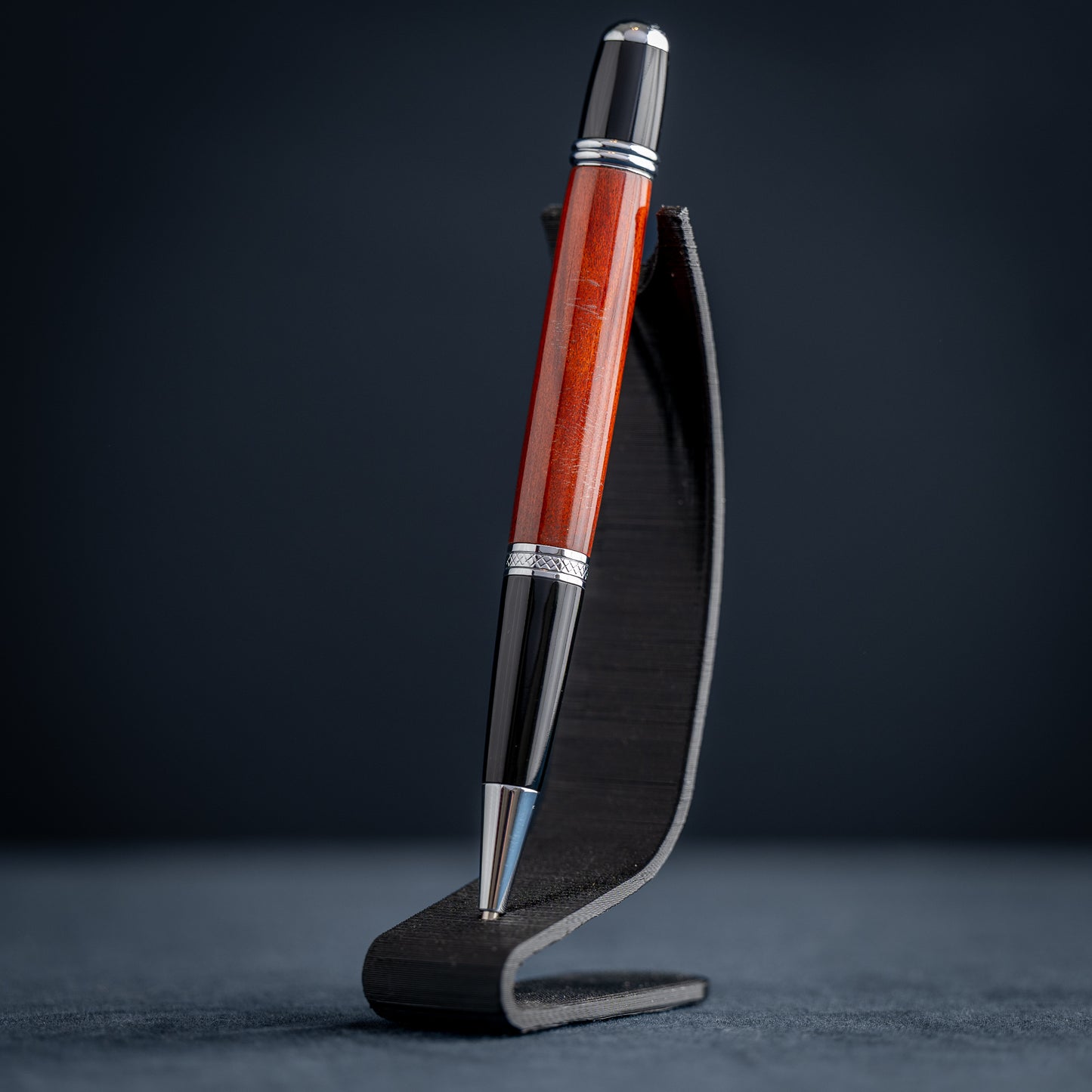 Handmade Redheart wooden ballpoint pen on a dark stand against a black background.