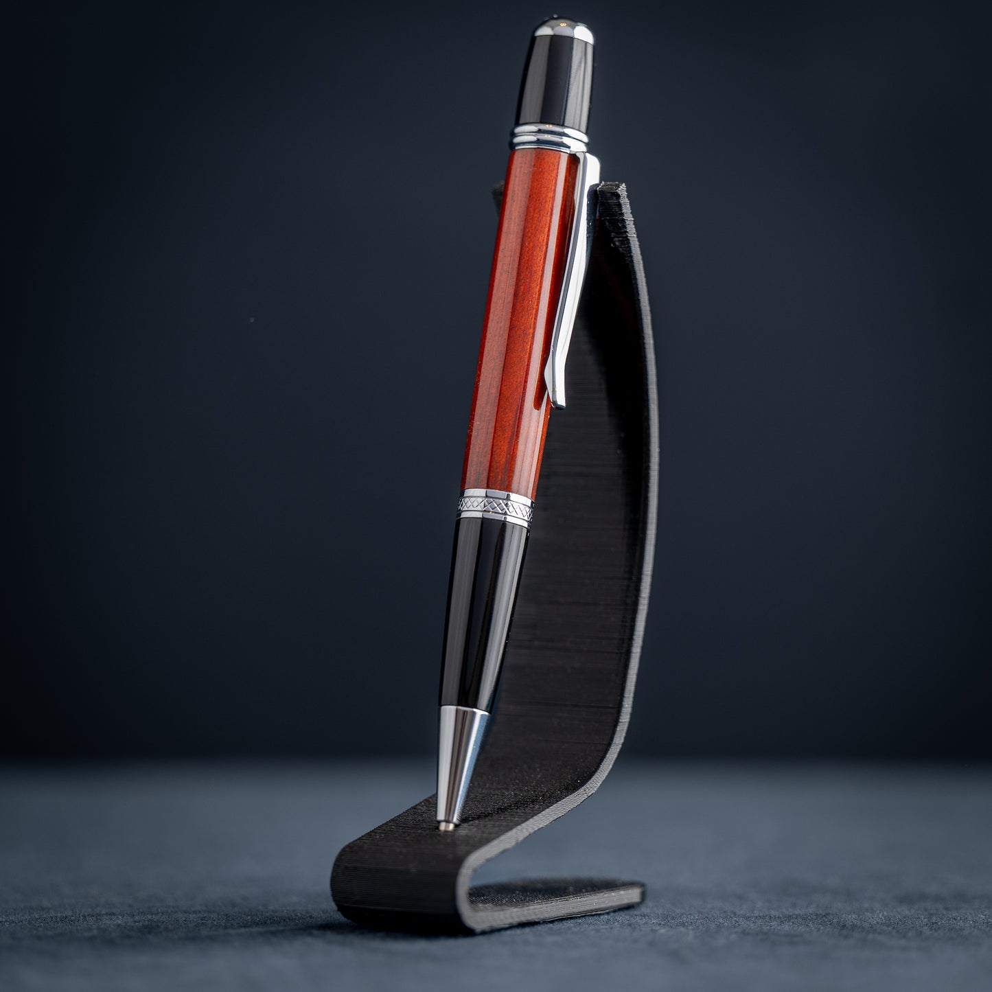 Handmade Redheart wooden ballpoint pen on a dark stand against a black background.