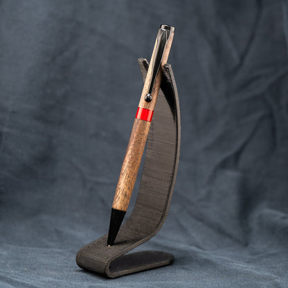 A handmade wooden twist pen made of Walnut wood and a custom red resin center band. The pen has black ink and black enamel plating.