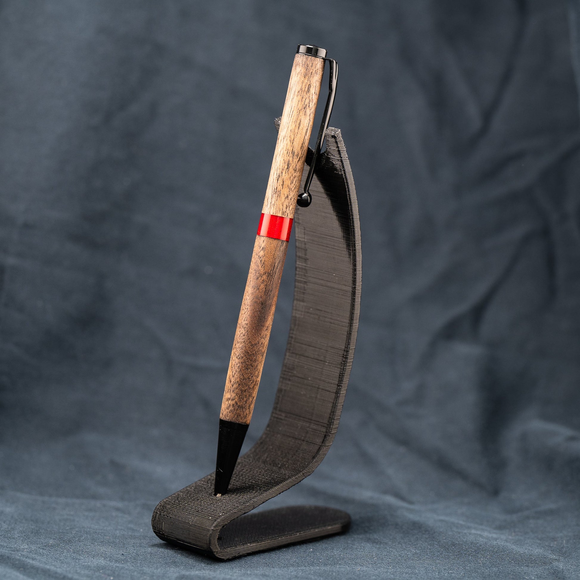 A handmade wooden twist pen made of Walnut wood and a custom red resin center band. The pen has black ink and black enamel plating.