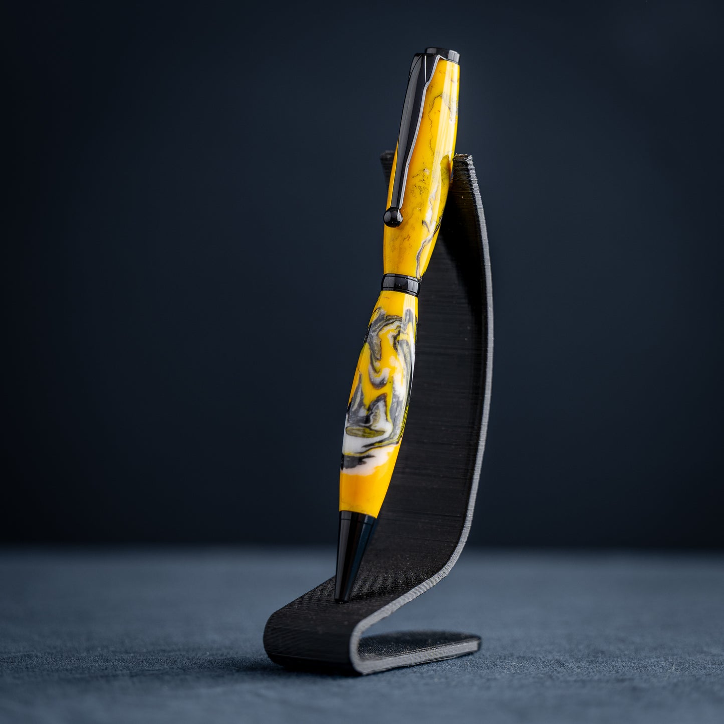 Handmade yellow, black, and white resin swirl ballpoint twist pen on a dark stand against a dark background.