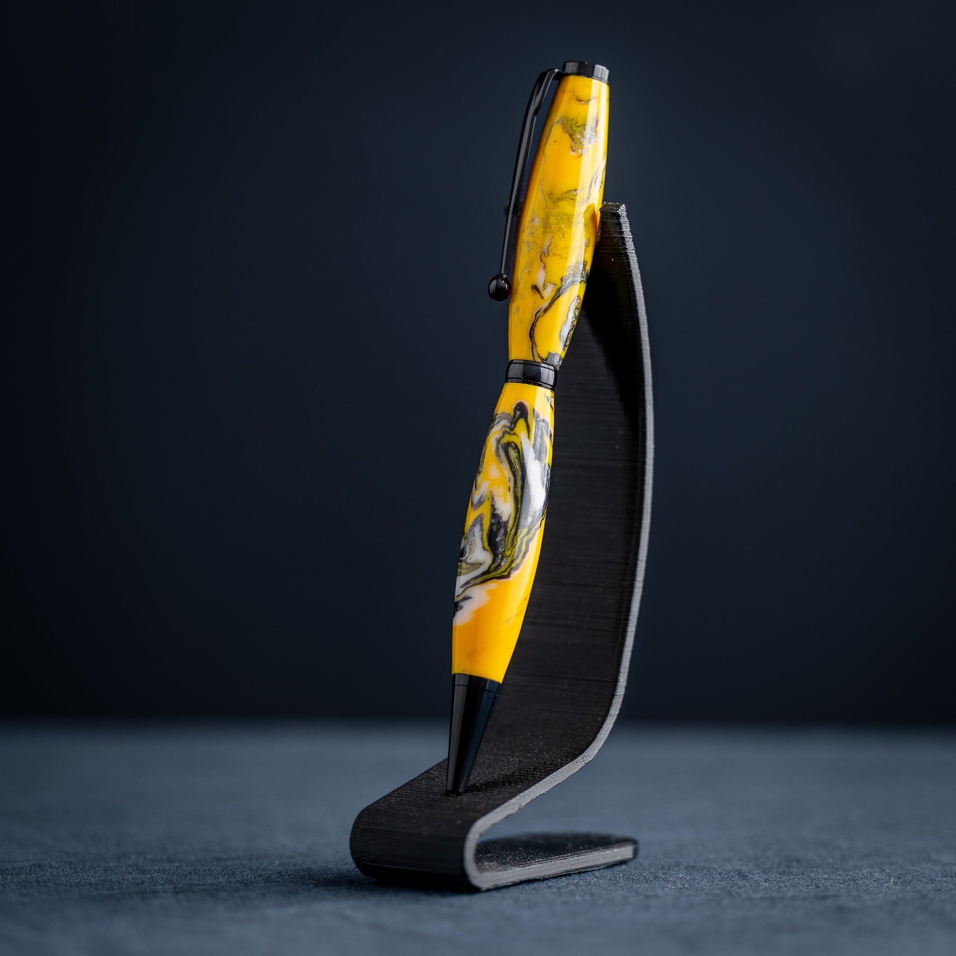 Handmade yellow, black, and white resin swirl ballpoint twist pen on a dark stand against a dark background.