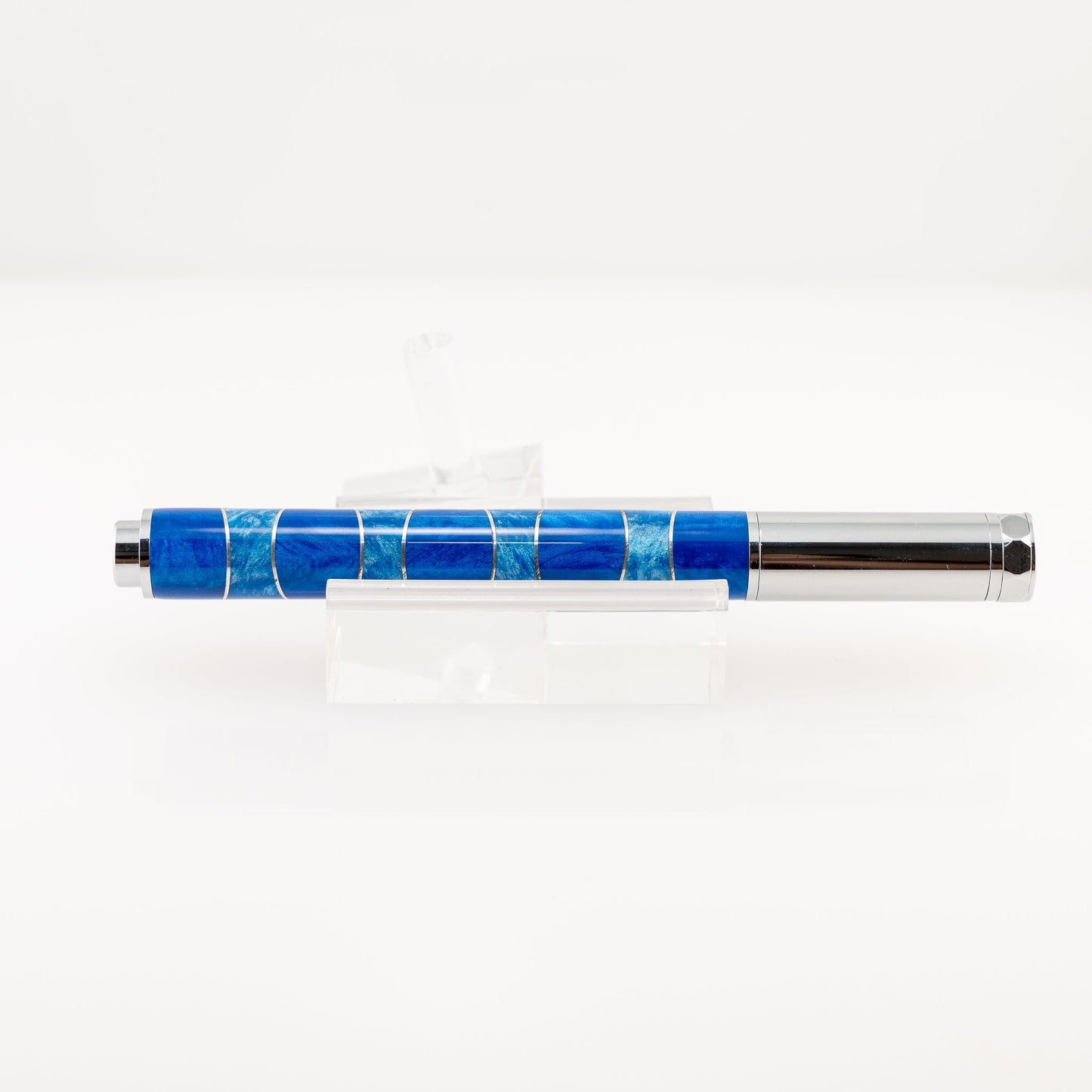 A handmade blue and light blue resin and aluminum fountain pen rests on a clear stand. The plating is chrome and it features a German Iridium nib.