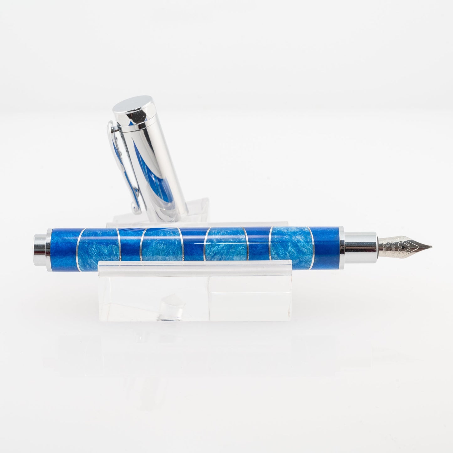 A handmade blue and light blue resin and aluminum fountain pen rests on a clear stand. The plating is chrome and it features a German Iridium nib.