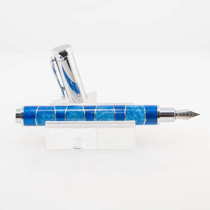 A handmade blue and light blue resin and aluminum fountain pen rests on a clear stand. The plating is chrome and it features a German Iridium nib.