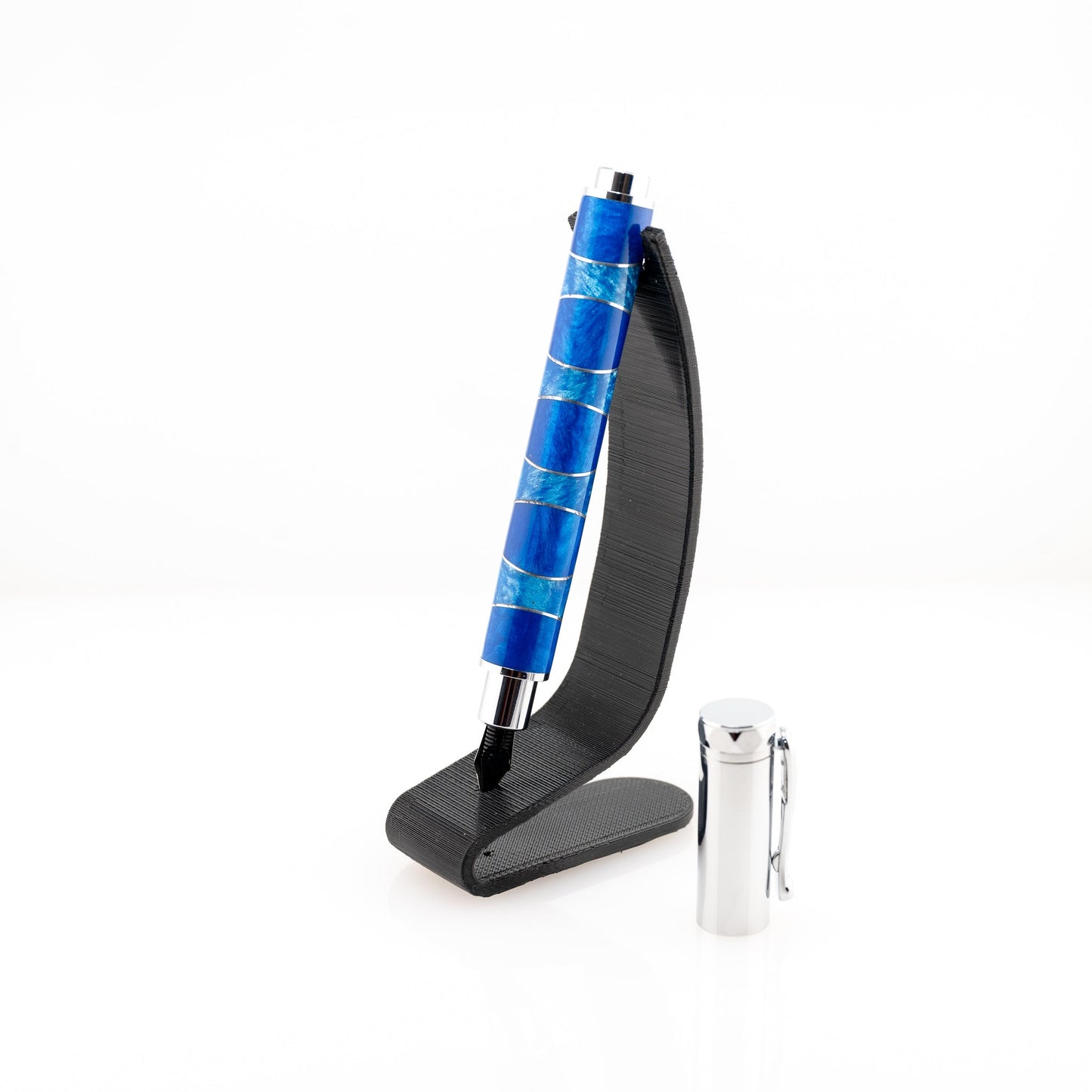 A handmade blue and light blue resin and aluminum fountain pen rests on a black stand. The plating is chrome and it features a German Iridium nib.